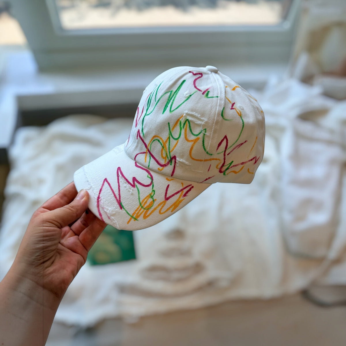 White cap with colorful abstract lines held by a hand in a room with a blurred background