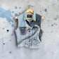 A sleeveless denim vest with a light wash and dark distressed panels, featuring a floral line-art graphic on the front pocket, hanging on a wooden hanger against a gray splattered background.