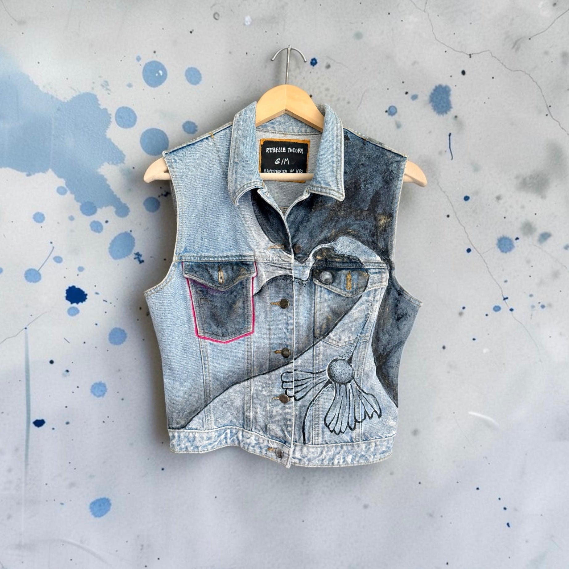 A sleeveless denim vest with a light wash and dark distressed panels, featuring a floral line-art graphic on the front pocket, hanging on a wooden hanger against a gray splattered background.