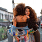 Two women embracing on a city street at sunset. The woman in front wears a black crop top and biker shorts, while the woman behind her has her back to the camera and is draped in a light blue pride  denim jacket covered in colorful graffiti-style paint splatters.