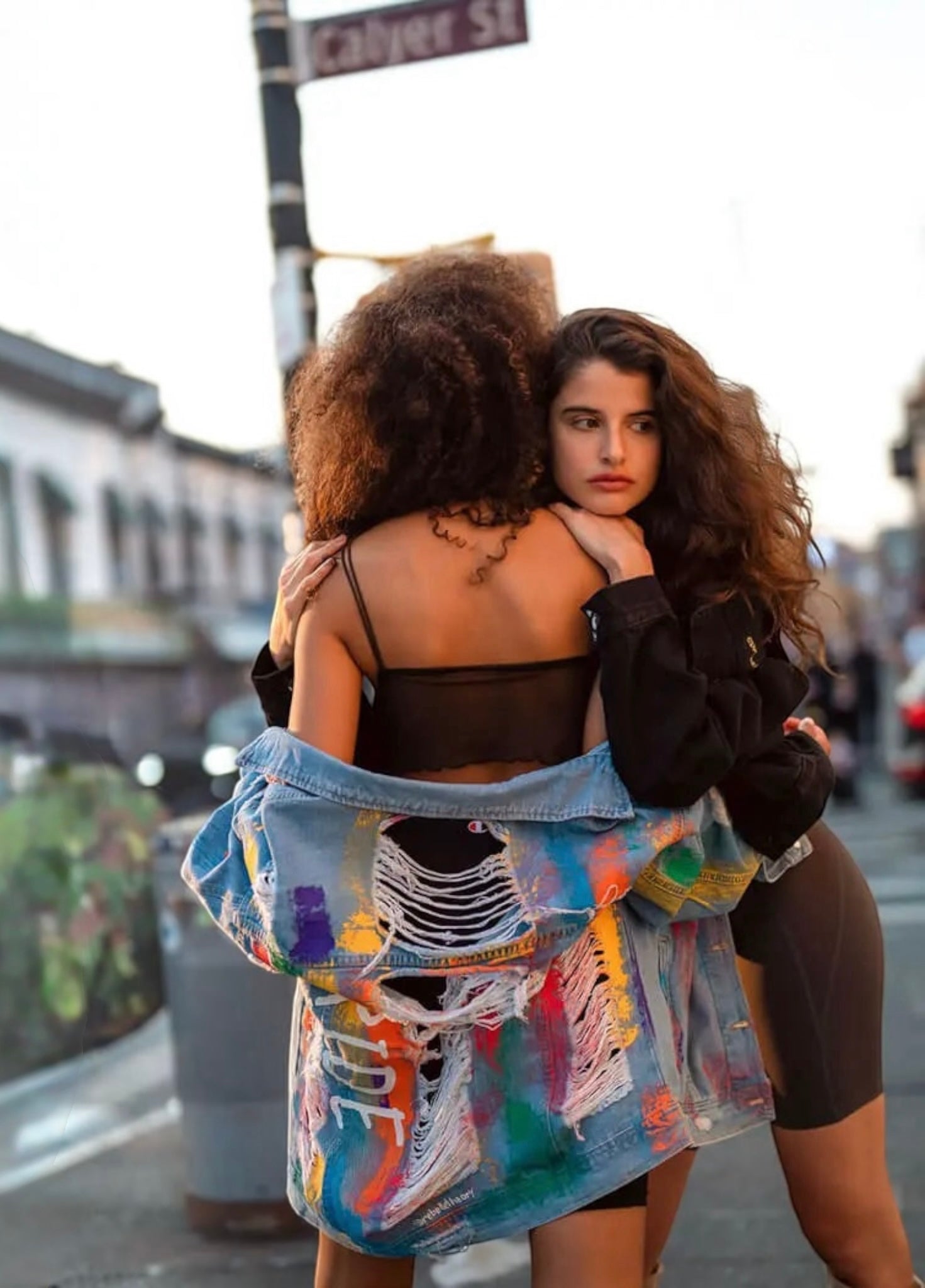 Two women embracing on a city street at sunset. The woman in front wears a black crop top and biker shorts, while the woman behind her has her back to the camera and is draped in a light blue pride  denim jacket covered in colorful graffiti-style paint splatters.