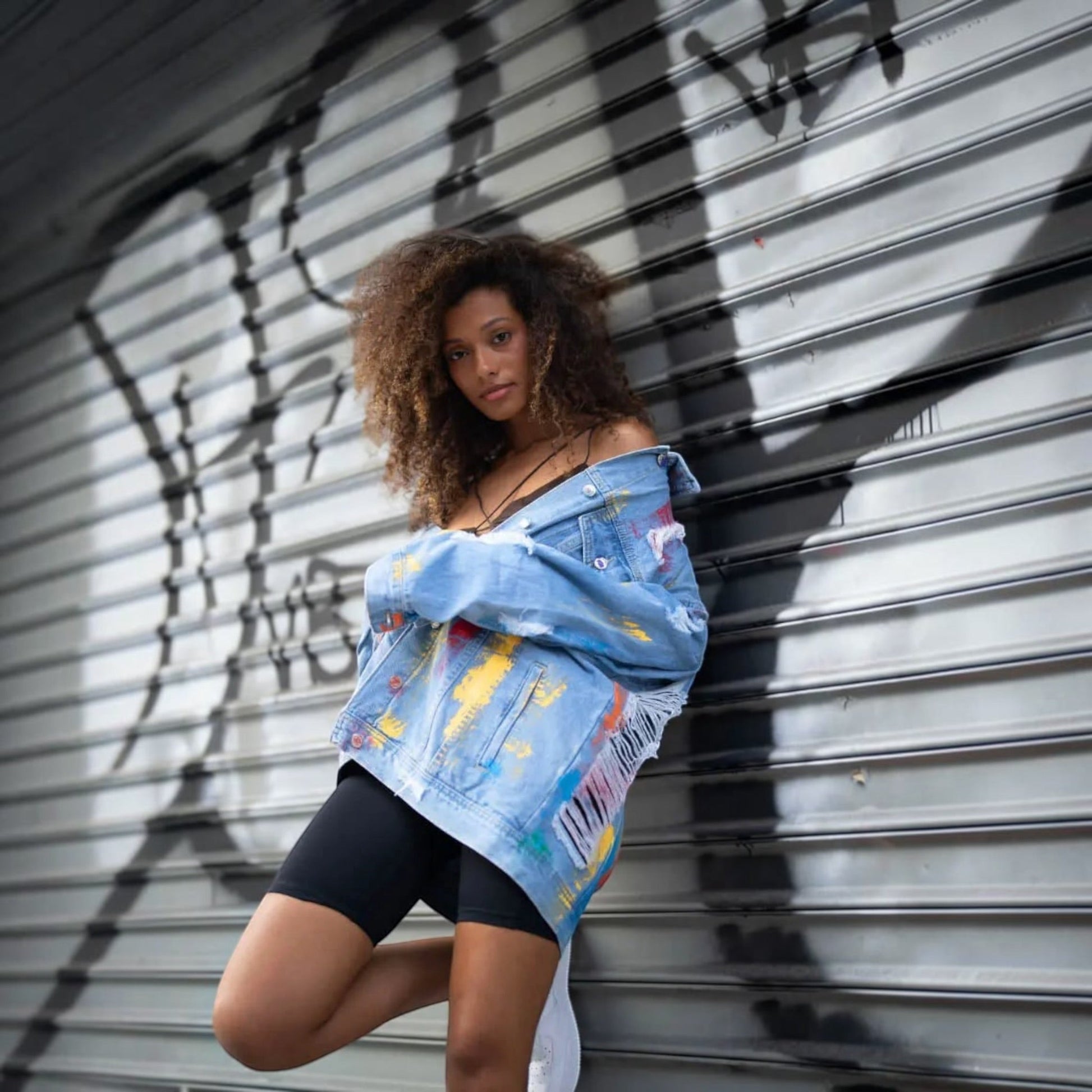 A woman posing in front of a gray metal shutter with black graffiti, wearing a light blue denim Pride jacket with colorful paint splatters and black fitted shorts