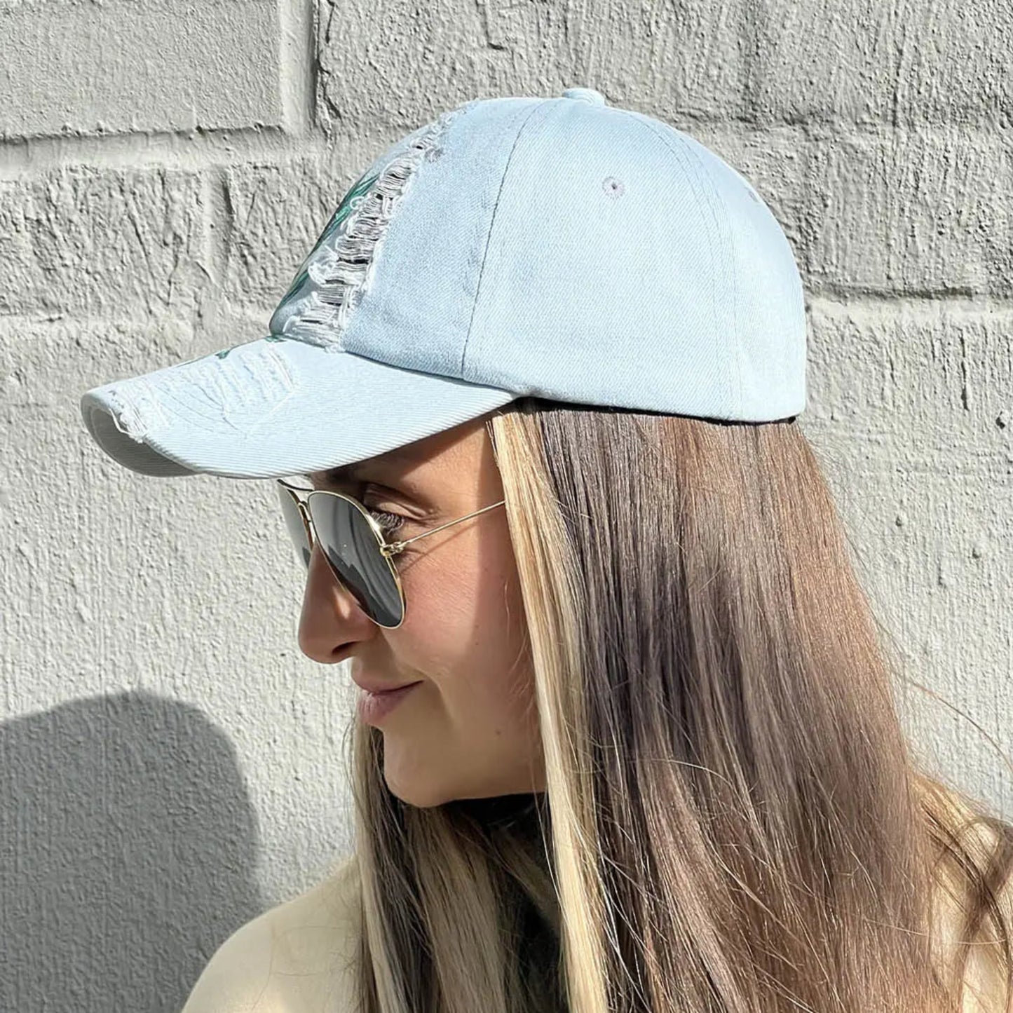 A person wearing sunglasses and a light blue/gray wash ripped denim baseball cap with an olive branch and three olives hand-painted on the front.