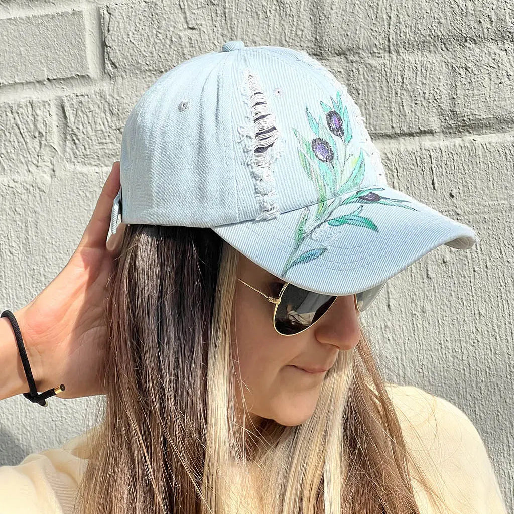 A person wearing sunglasses and a light blue/gray wash ripped denim baseball cap with an olive branch and three olives hand-painted on the front.