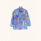 A light blue denim jacket with colorful paint splatter and patch designs, hanging on a wooden hanger against a solid cream background.
