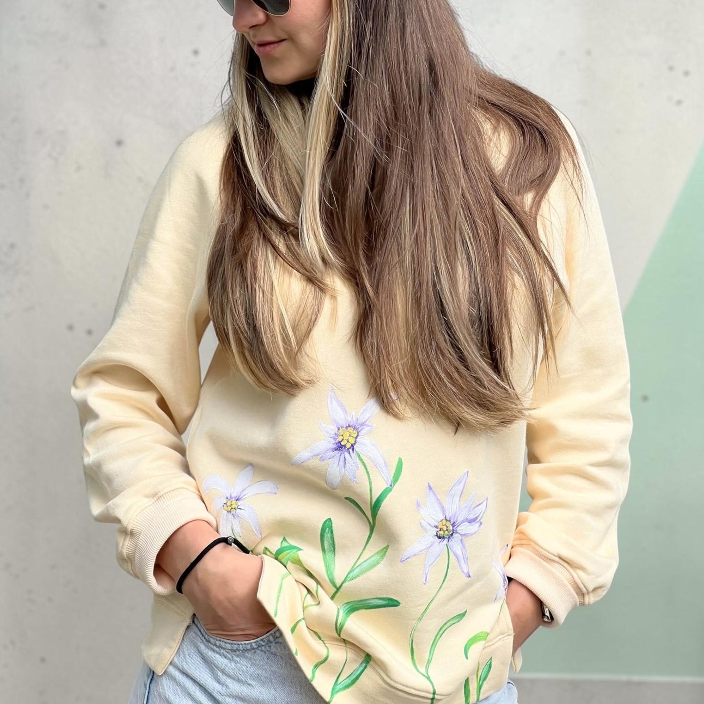 A woman wearing a cream-colored crewneck sweatshirt with a green and purple floral print, paired with light blue jeans.