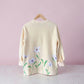 A cream-colored long-sleeve sweatshirt with a floral print of light purple flowers and green stems, hanging on a wooden hanger against a soft pink background.