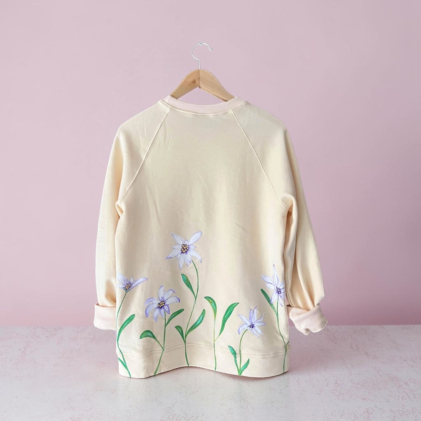 A cream-colored long-sleeve sweatshirt with a floral print of light purple flowers and green stems, hanging on a wooden hanger against a soft pink background.