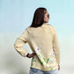 A woman shown from the back wearing a light beige sweatshirt with a floral print of white flowers and green leaves along the lower hem, paired with light blue jeans against a light blue gradient background.