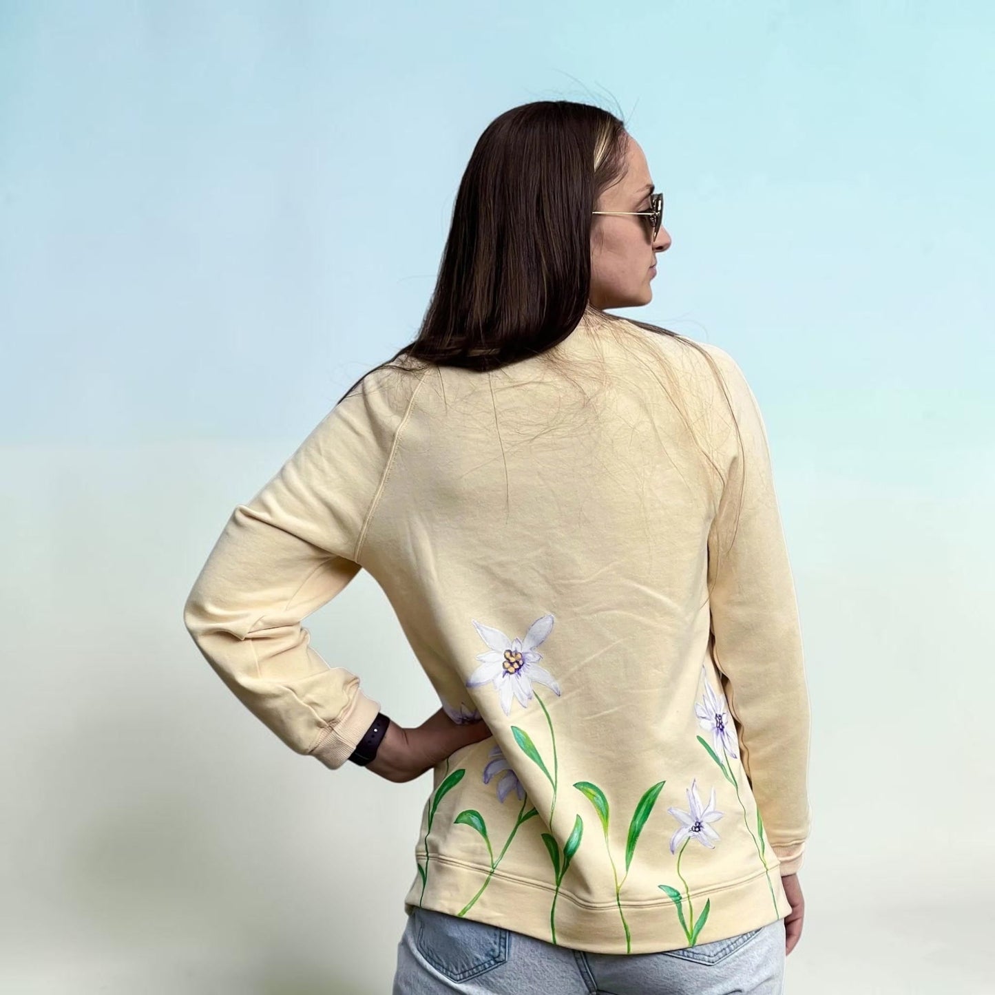 A woman shown from the back wearing a light beige sweatshirt with a floral print of white flowers and green leaves along the lower hem, paired with light blue jeans against a light blue gradient background.