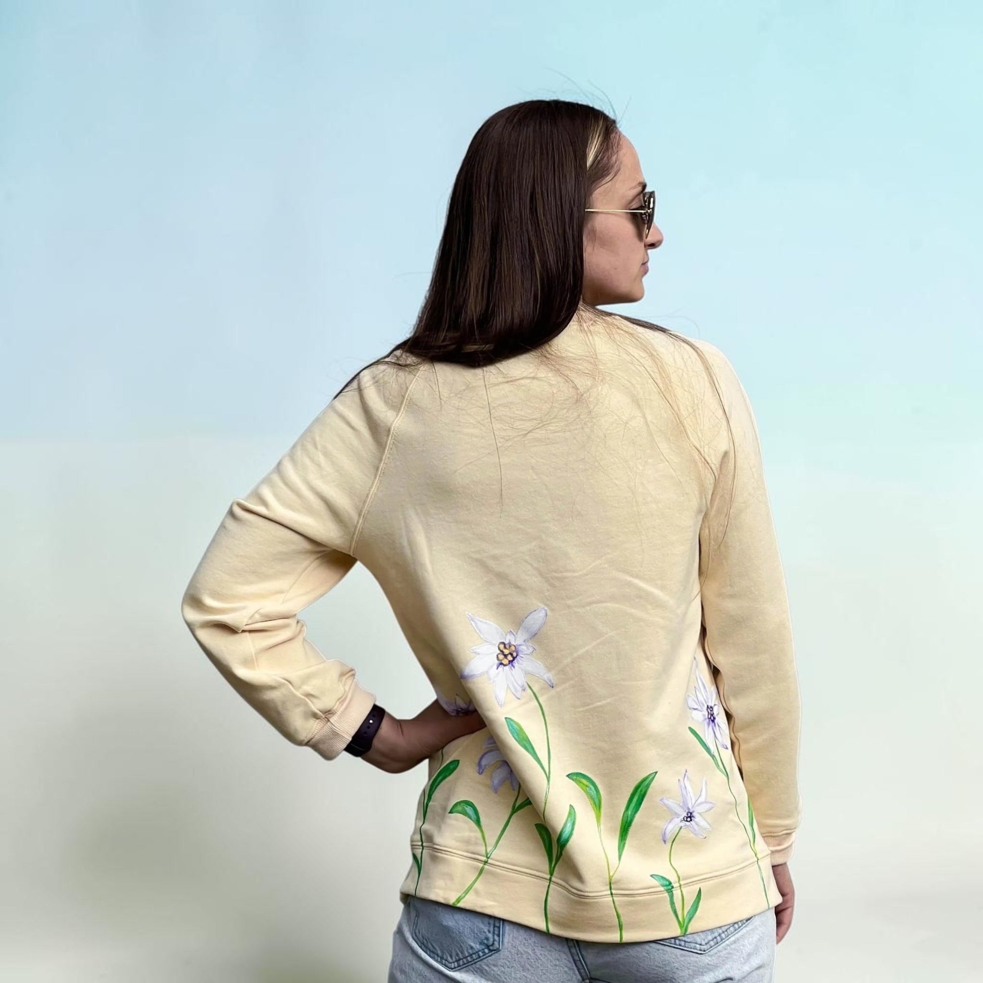 A woman shown from the back wearing a light beige sweatshirt with a floral print of white flowers and green leaves along the lower hem, paired with light blue jeans against a light blue gradient background.