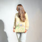 A woman with long light brown hair is wearing a pale yellow sweatshirt with a green and purple floral pattern on the lower back, paired with light gray jeans, set against a plain white wall.