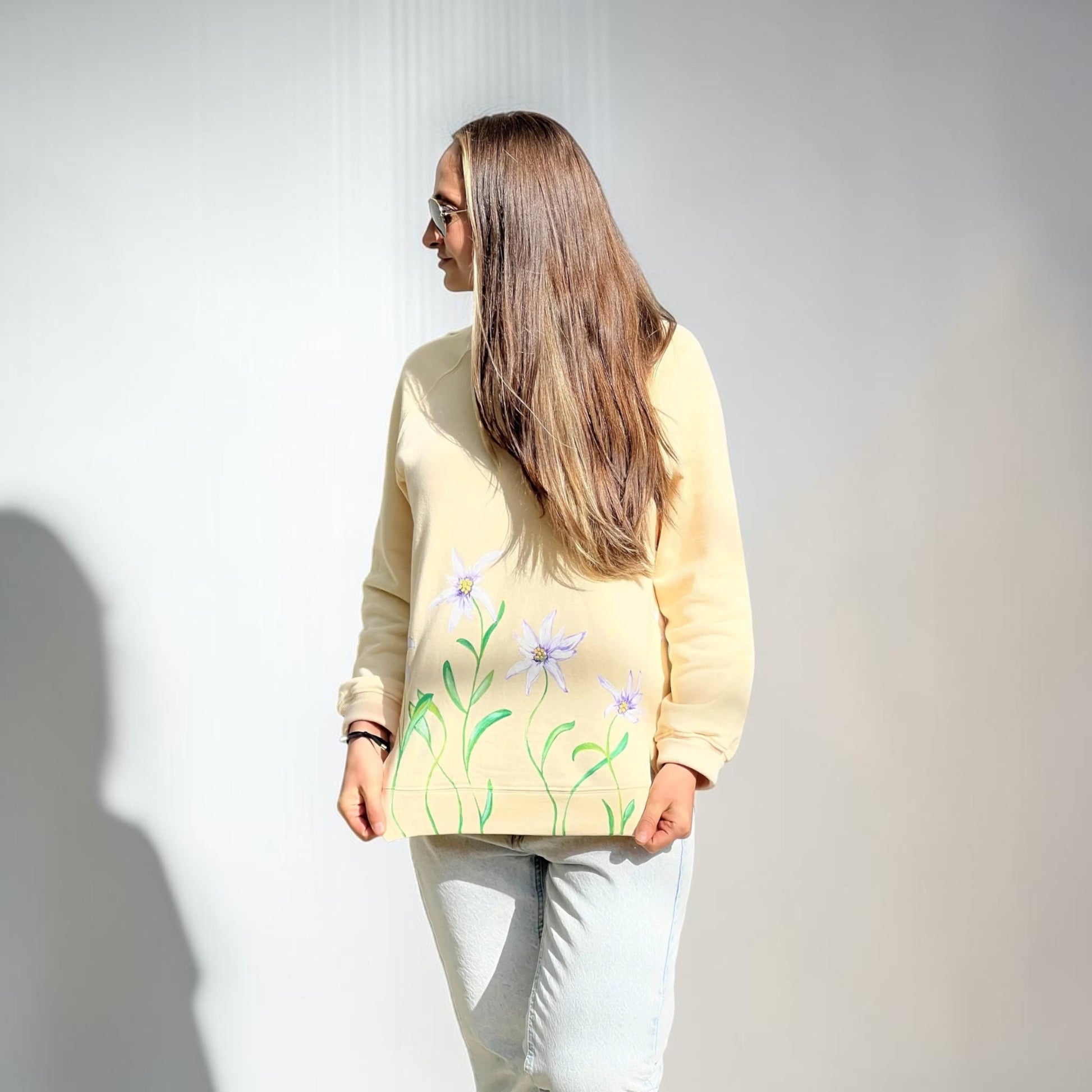 A woman with long light brown hair is wearing a pale yellow sweatshirt with a green and purple floral pattern on the lower back, paired with light gray jeans, set against a plain white wall.