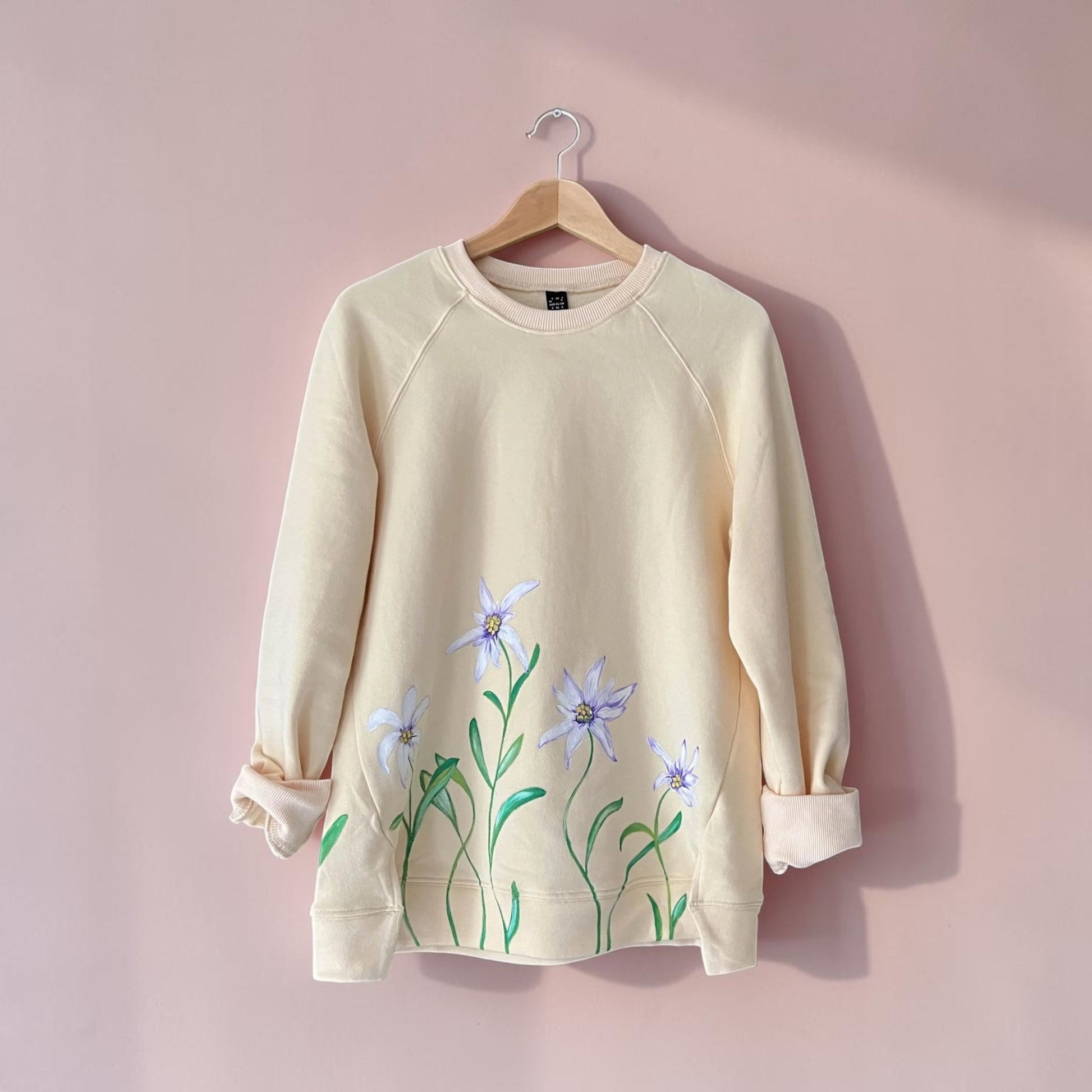 A cream-colored long-sleeve sweatshirt with a floral print of light purple and green flowers, hanging on a wooden hanger against a soft pink wall.