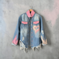 A light blue distressed denim jacket with pink accents, hanging on a wooden hanger against a textured gray concrete wall. The jacket features a pink collar and pocket flaps, a large pink heart graphic on the left chest, and frayed details along the hem and cuffs.
