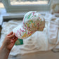White cap with colorful abstract lines held by a hand in a room with a blurred background