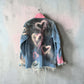 A distressed denim jacket hanging on a metal hanger against a textured concrete wall. The back of the jacket is faded blue with hand-painted pink and white heart designs, featuring raw, frayed edges and ripped details near the hem and cuffs.