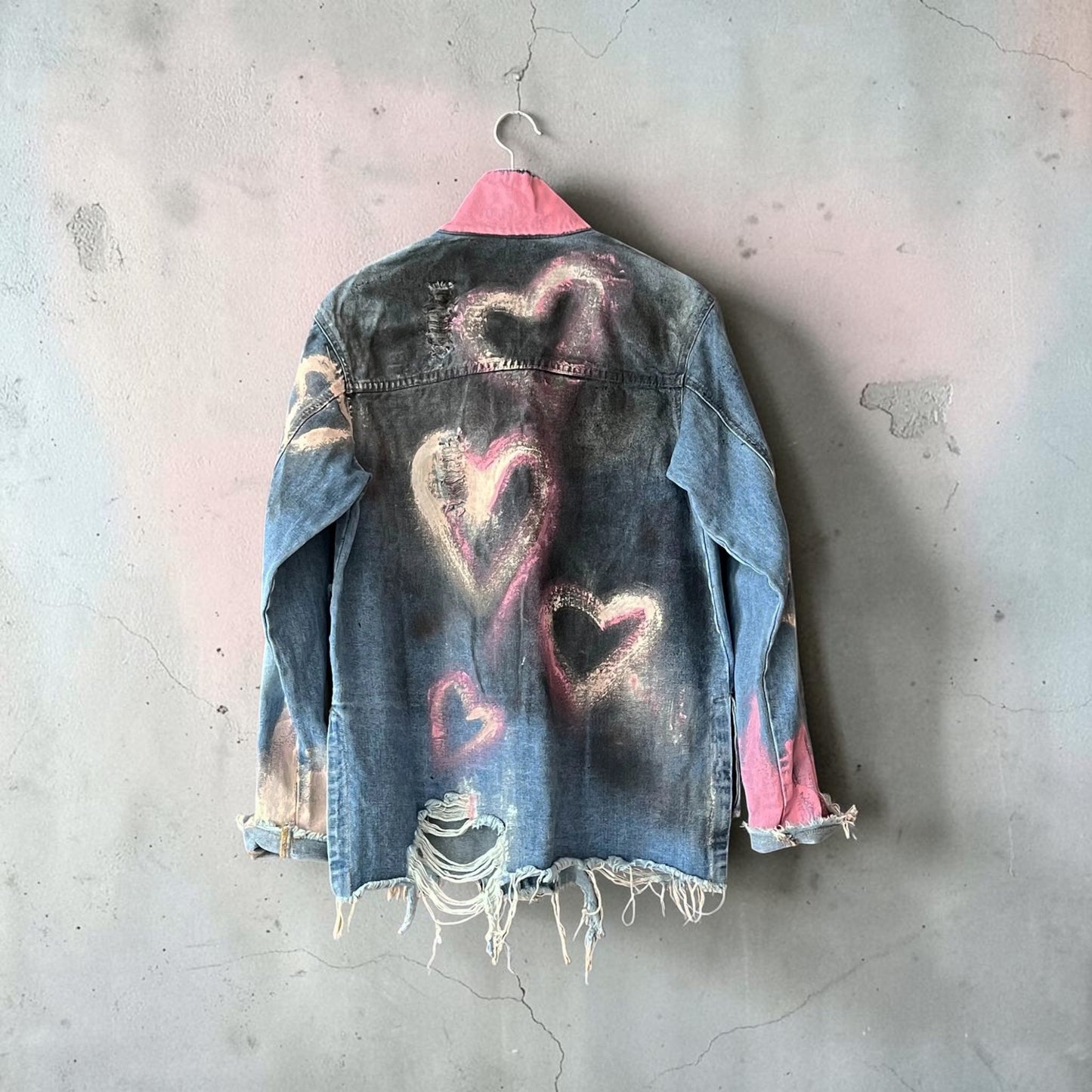 A distressed denim jacket hanging on a metal hanger against a textured concrete wall. The back of the jacket is faded blue with hand-painted pink and white heart designs, featuring raw, frayed edges and ripped details near the hem and cuffs.