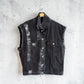 A sleeveless black denim vest with a distressed, faded wash and silver-toned metal buttons, hanging on a wooden hanger against a textured gray concrete wall.