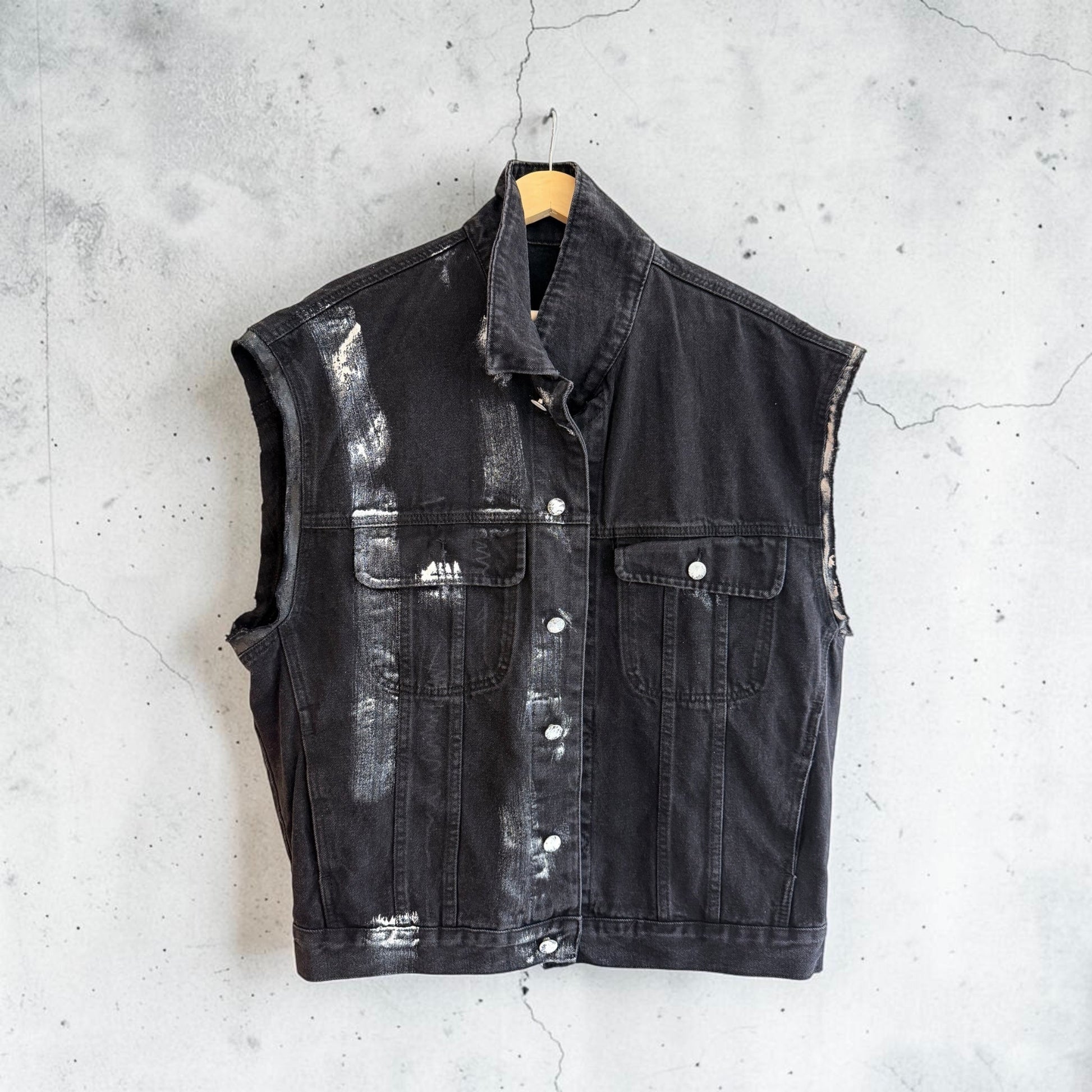 A sleeveless black denim vest with a distressed, faded wash and silver-toned metal buttons, hanging on a wooden hanger against a textured gray concrete wall.