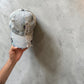 A light-wash distressed denim baseball cap with olives painted , held by a hand against a gray concrete floor.