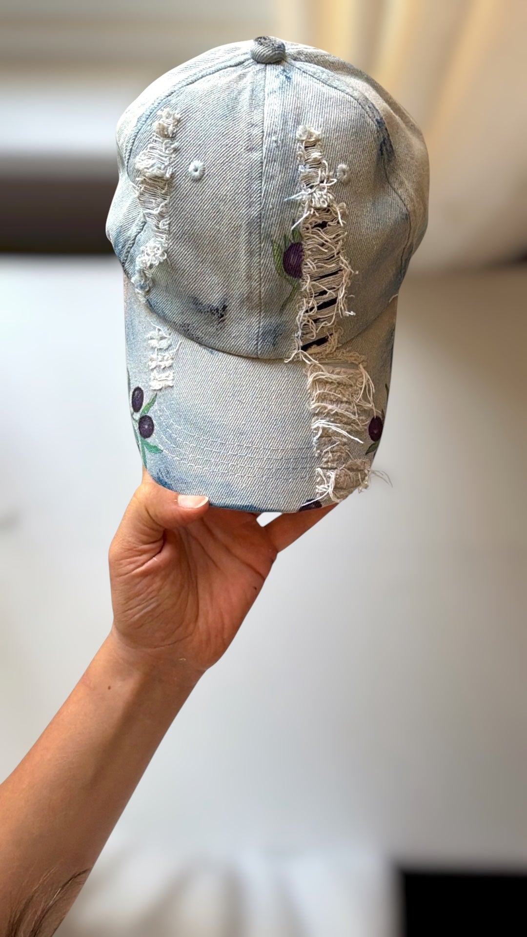 A light blue distressed denim baseball cap held up by a hand, featuring ripped details and frayed edges with hand painted olives