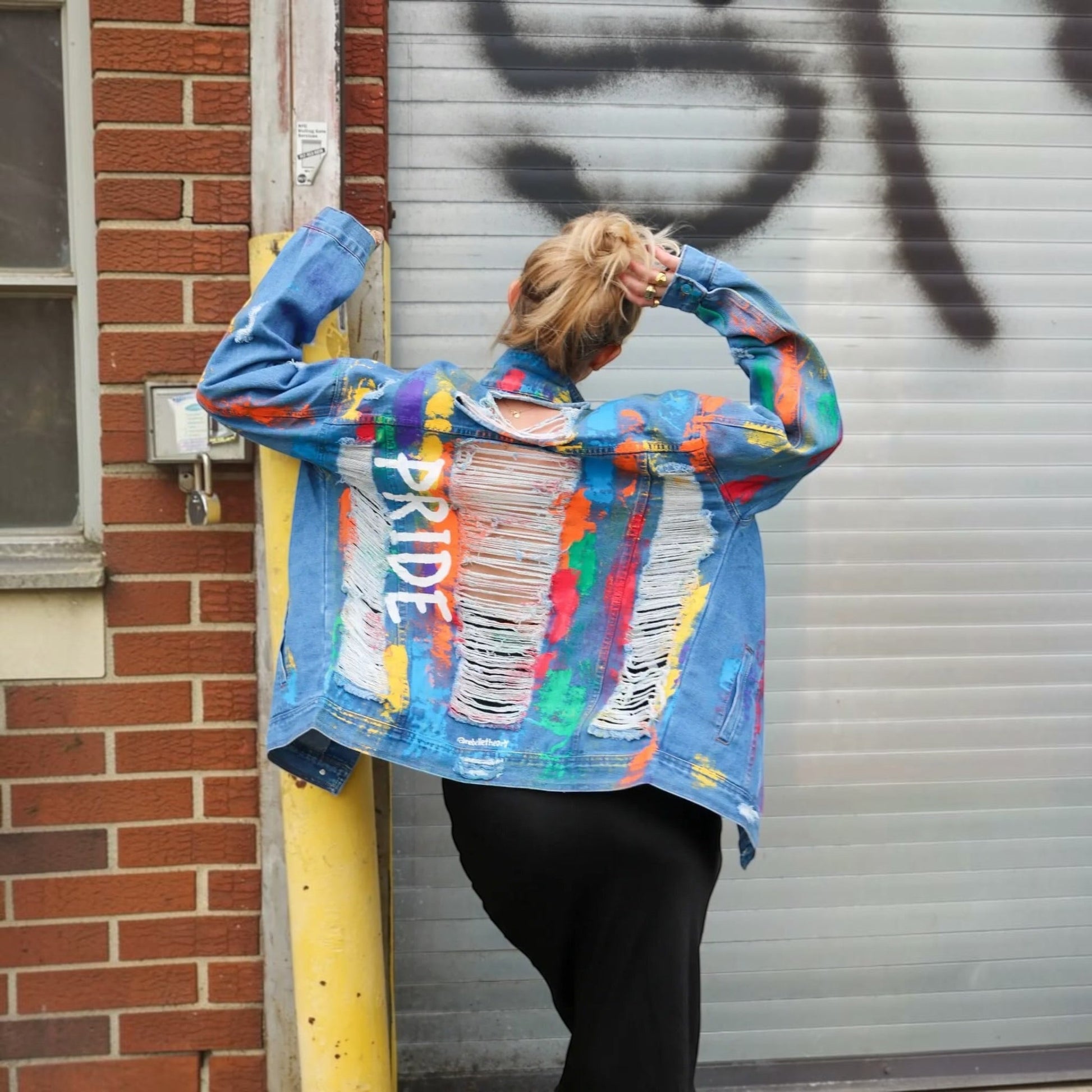 A person stands with their back to the camera wearing a blue denim jacket covered in colorful paint splatters and ripped-paper effects, with the word 'PRIDE' printed vertically on the back. They are posing in front of a brick wall, a yellow utility pole, and a gray metal garage door with graffiti.
