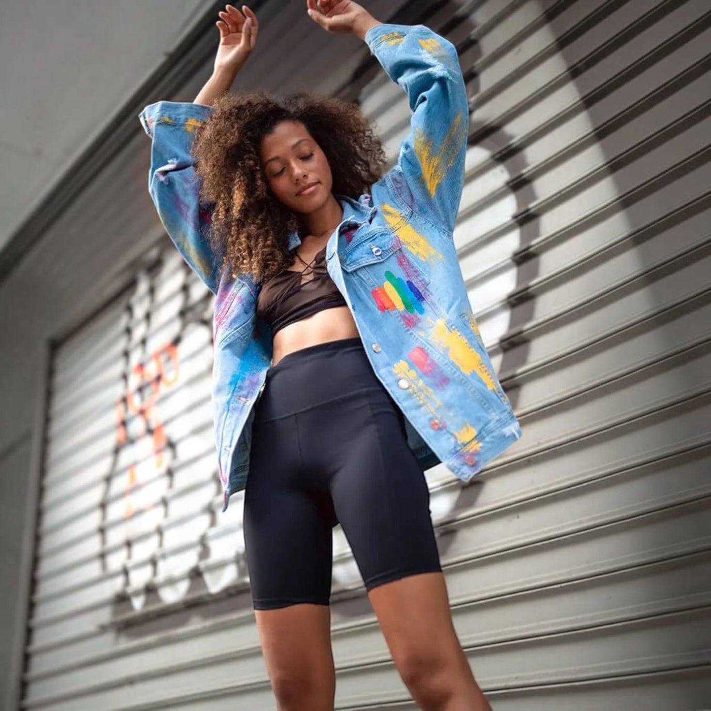 A woman posing in front of a gray metal shutter, wearing a light blue  Pride denim jacket with colorful paint splatters, a black sports bra, and black biker shorts.