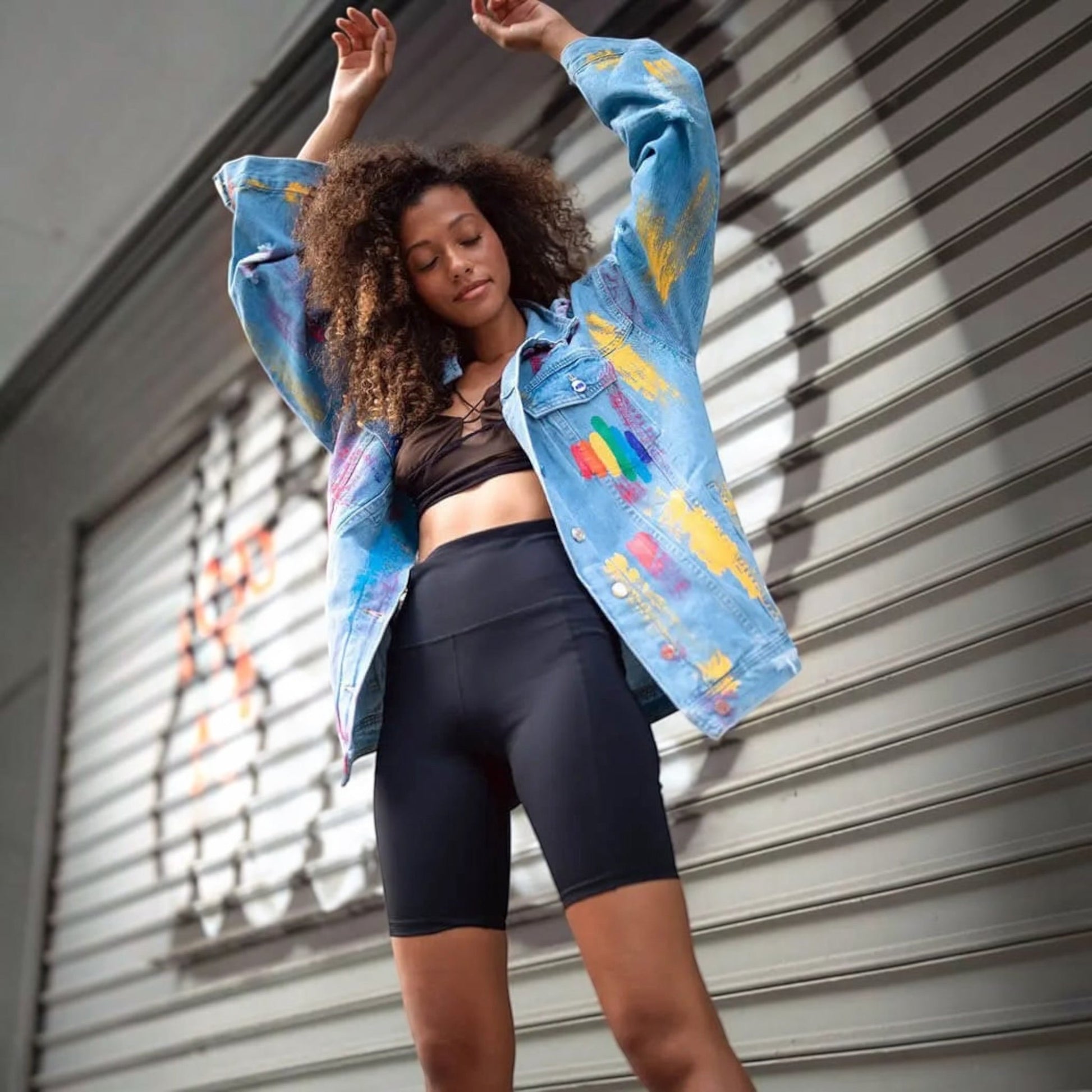 A woman posing in front of a gray metal shutter, wearing a light blue  Pride denim jacket with colorful paint splatters, a black sports bra, and black biker shorts.