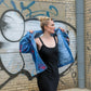 A woman stands against a graffiti-covered brick wall, holding open a blue denim jacket with red paint splatters over her shoulders while wearing a black sleeveless dress.