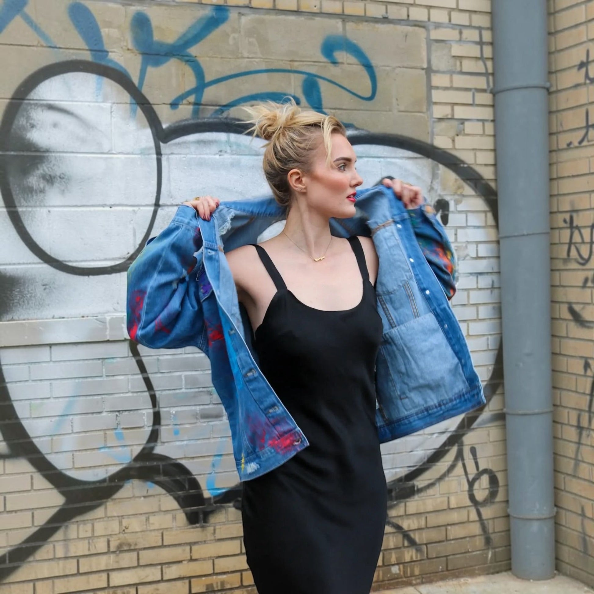 A woman stands against a graffiti-covered brick wall, holding open a blue denim jacket with red paint splatters over her shoulders while wearing a black sleeveless dress.