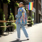 A person walking outdoors on a city sidewalk wearing a light blue denim jacket with colorful graffiti-style paint patches and the word 'PRIDE' on the back, paired with light blue jeans and white sneakers. A rainbow flag hangs in the background.