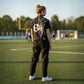 A woman stands on a green soccer field wearing a black denim vest with a soccer ball graphic on the back, a black t-shirt, black cargo pants, and white sneakers.