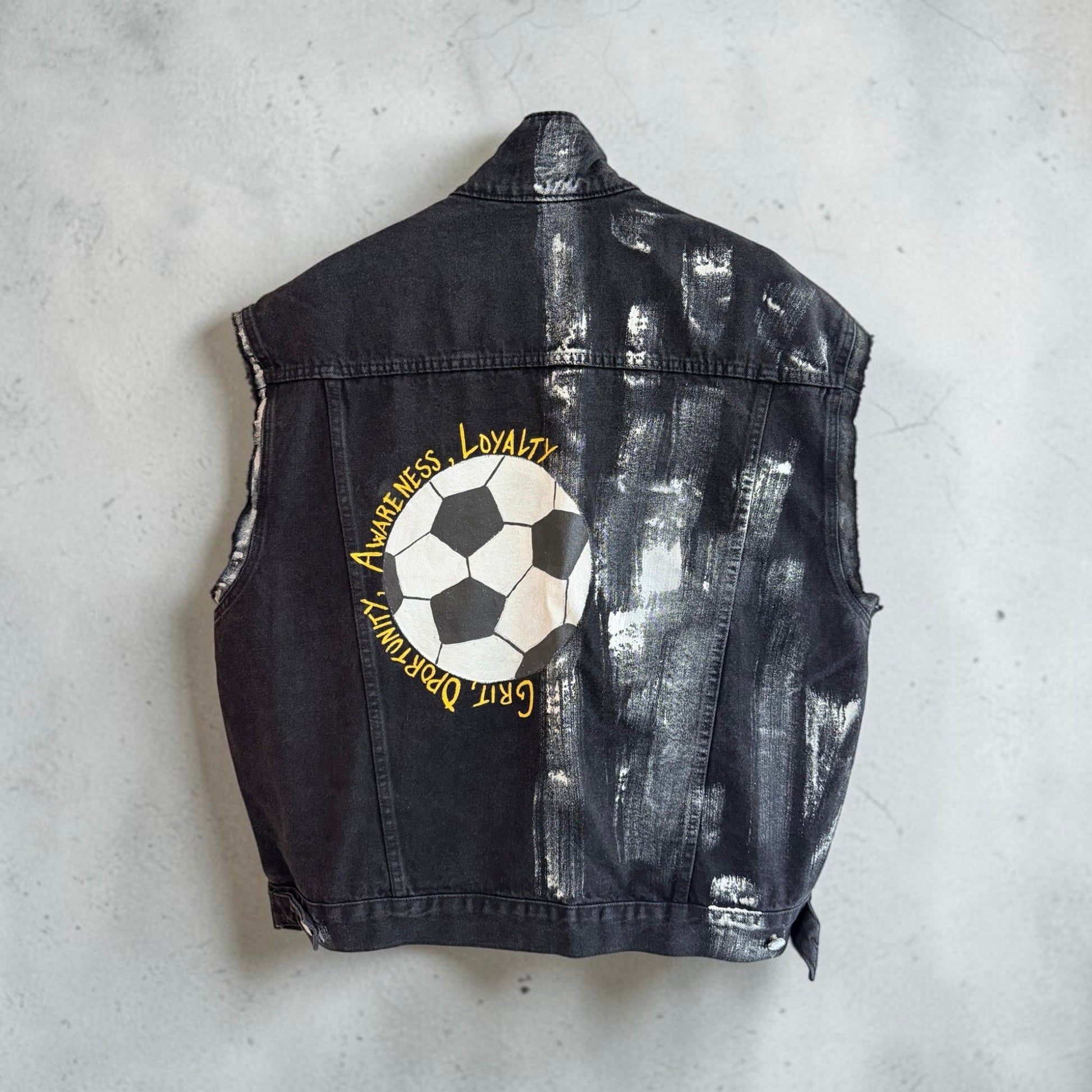 A black washed denim vest shown from the back, featuring a large graphic print of a soccer ball with the words 'Awareness, Loyalty' and 'Gert D'Armony' in yellow text, laid flat on a light gray concrete surface.