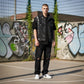 A man stands on an outdoor basketball court wearing a black denim vest over a black t-shirt, black ripped jeans, and white sneakers. The background features a graffiti-covered wall and a chain-link fence.