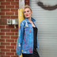 A woman wearing a light blue distressed denim jacket with colorful paint splatters, layered over a black top, standing in front of a brick wall and a gray metal shutter.