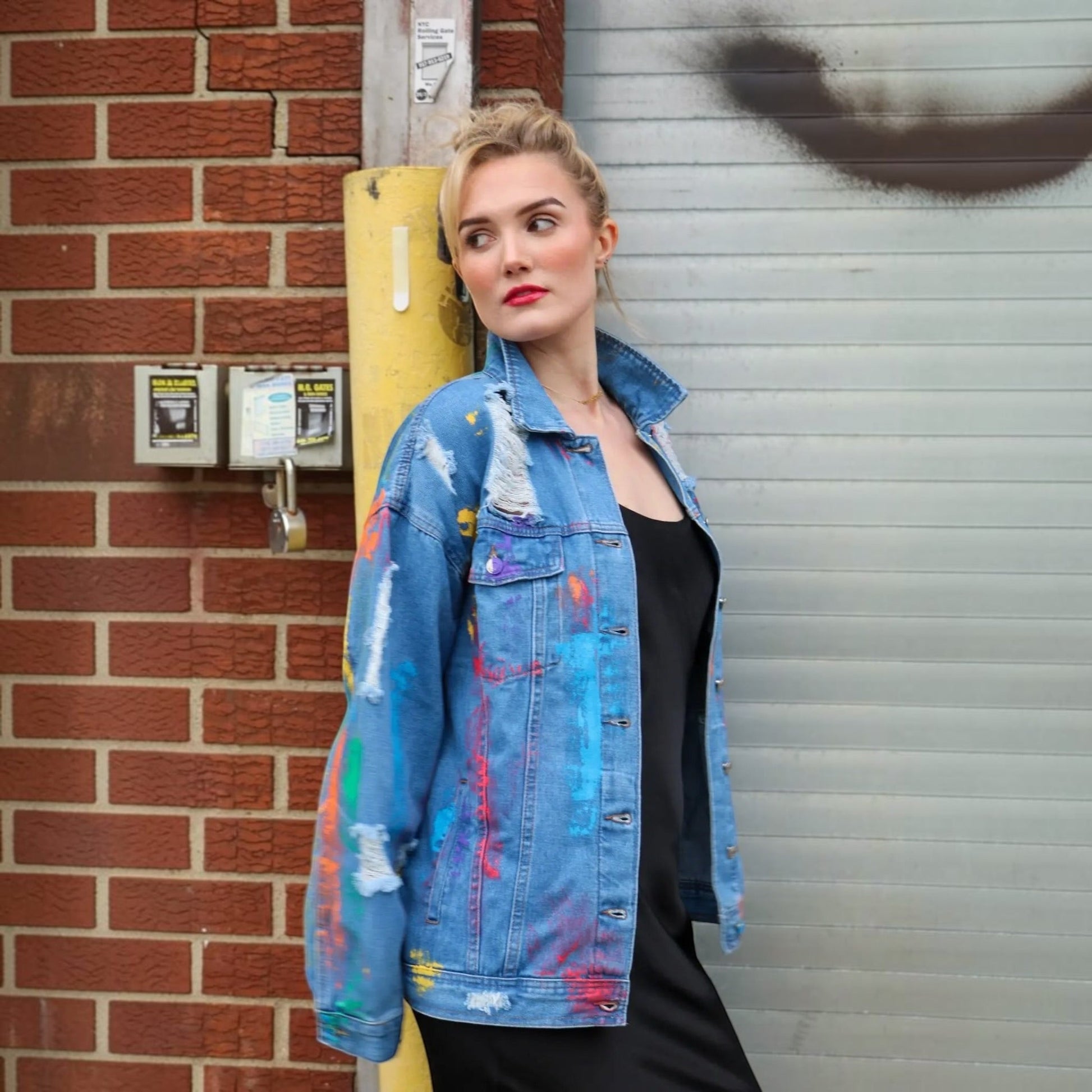 A woman wearing a light blue distressed denim jacket with colorful paint splatters, layered over a black top, standing in front of a brick wall and a gray metal shutter.